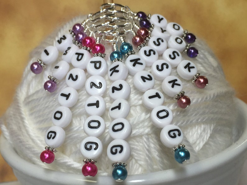 May include: White plastic stitch markers with black lettering, including numbers and letters, with colorful beads attached to each marker. The markers are arranged in a circle on a white yarn ball.