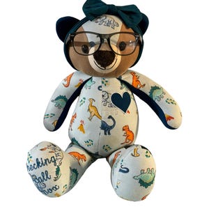 May include: A plush teddy bear with a blue bow and glasses, wearing a blue and white fabric with a dinosaur print. The bear has a heart-shaped patch on its chest and the words "checking ball now" embroidered on one foot.