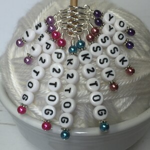 May include: White plastic beads with black letters spelling "K2tog, K2tog, P2tog, P2tog, K2tog, K2tog, YOS, YOS" and numbers "1, 0, 0, 6, 1, 0, 0, 6" on a white yarn ball. Each bead has a silver ring and a colorful bead.