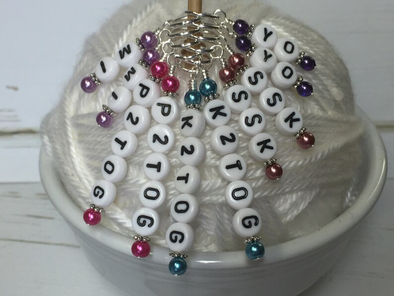 May include: White plastic beads with black letters spelling "K2tog, K2tog, P2tog, P2tog, K2tog, K2tog, YOS, YOS" and numbers "1, 0, 0, 6, 1, 0, 0, 6" on a white yarn ball. Each bead has a silver ring and a colorful bead.