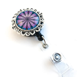 May include: A silver retractable badge holder with a black strap and a round, ornate, silver frame with a pink and blue mandala design.