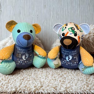 May include: Two stuffed teddy bears made from patterned fabric. Each bear features a dark blue torso embroidered with the name Desmond Vaughn Cooper, birth details including 8 lbs 4 ozs, 19.5 ins, and the date 05-21-2024, alongside two small footprints.
