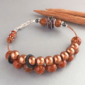 May include: A copper-toned bracelet with a silver clasp and a variety of beads, including a striped glass bead, copper-toned metal beads, and brown speckled beads.