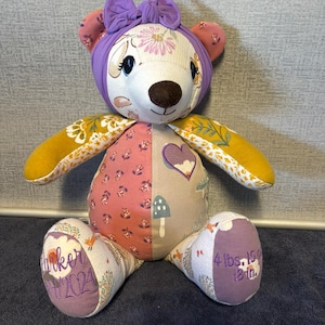 May include: A handmade teddy bear with a purple bow, floral accents, and embroidered details. The bear's body is a patchwork of patterned fabrics in pink, yellow, and purple. The bear's foot reads "4 lbs. 15 oz. 18 in."