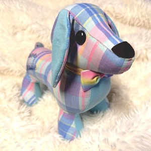 May include: A plush dachshund toy made of light blue, purple, and yellow plaid fabric. The dog features solid light blue ears, a black nose, large black eyes, and a small rainbow-striped bow tie around its neck. The word "love" is visible on its side.
