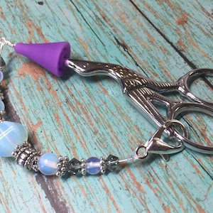 May include: A silver pair of scissors with a purple silicone tip attached to a silver chain with blue and silver beads.