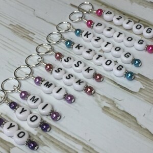May include: A set of eight white bead keychains with black letter beads spelling out the words "YO", "MI", "SSK", "K2TOG", "P2TOG". Each keychain has a silver ring and colorful beads.