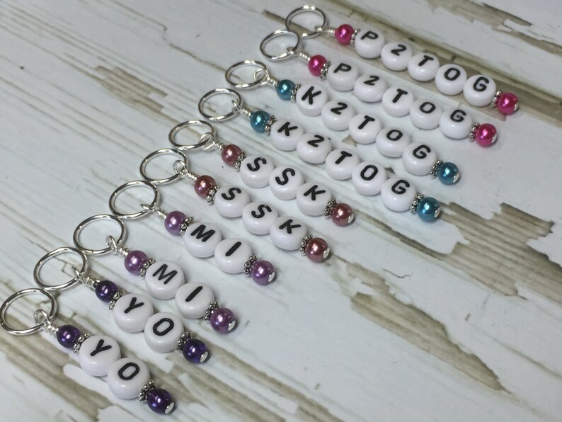 May include: A set of eight white bead keychains with black letter beads spelling out the words "YO", "MI", "SSK", "K2TOG", "P2TOG". Each keychain has a silver ring and colorful beads.