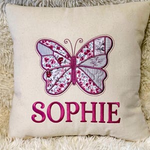 May include: A square off-white throw pillow featuring an embroidered butterfly design in shades of pink, grey, and white with floral and striped patterns. The name SOPHIE is embroidered in bold pink capital letters centered below the butterfly.