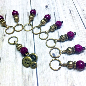 May include: Set of eight stitch markers with antique gold rings and purple beads. Each marker features a decorative bead with a textured surface. One marker has a charm with three buttons.
