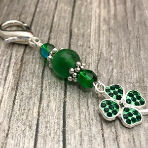 May include: A green four-leaf clover charm with silver accents hangs from a silver clip with green and clear glass beads.