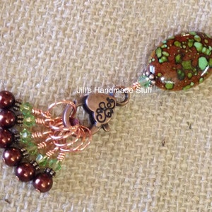 May include: A brown and green speckled oval bead with copper wire and green glass beads attached to a copper charm with the text "Jill's Handmade Stuff" and a heart design. The charm is attached to a set of copper rings with brown beads.