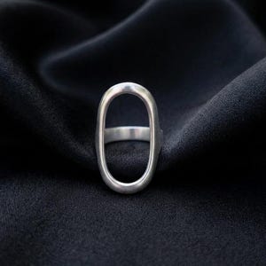 May include: A silver ring with an elongated oval design. The ring has a smooth, polished finish and a minimalist aesthetic. The ring is set against a black background.