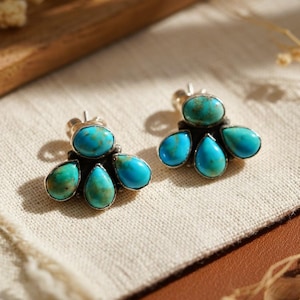 May include: A pair of turquoise stud earrings. Each earring has a cluster of turquoise stones set in silver. The stones are a vibrant blue-green colour, with a teardrop shape. The earrings are displayed on a neutral-coloured fabric.