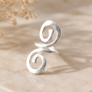 May include: A silver-toned ring featuring a double spiral design with a hammered, textured finish. The metal band curves into two distinct circular coils, one positioned above the other, creating an open, continuous shape with a reflective surface.