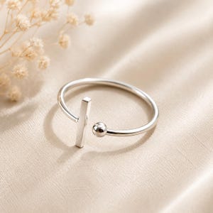 May include: A silver-toned open ring featuring a thin, polished band. One end of the band terminates in a small, vertical rectangular bar, while the opposite end features a smooth, spherical bead. The design is simple, metallic, and reflective.