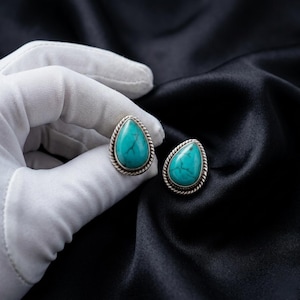 May include: A pair of teardrop-shaped turquoise earrings with a silver-colored rope-like border. The earrings are displayed on a black satin fabric. The turquoise stones have natural veining.