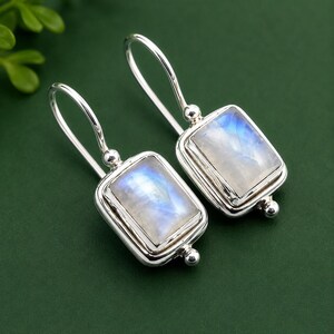 May include: A pair of drop earrings featuring rectangular moonstone cabochons set in polished silver frames. Each earring has a curved silver ear wire and a small silver bead accent at the base of the setting, showcasing the iridescent blue sheen.