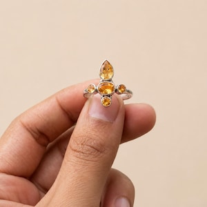 May include: A silver ring featuring a central oval-cut yellow citrine gemstone, flanked by two smaller round-cut citrine stones on the sides and one pear-cut citrine stone positioned at the top, all set in a polished silver metal band.