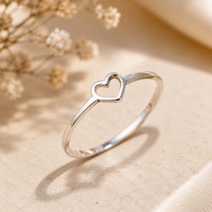 May include: A thin, polished silver-tone ring featuring a delicate, open-work heart shape centered on the band. The metal surface is smooth and reflective, catching the light to create a bright, metallic sheen across the circular ring structure.