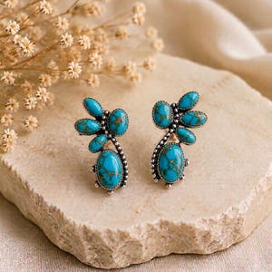 May include: A pair of stud earrings featuring bright turquoise stones with brown matrix patterns set in silver metal. Each earring has a cluster of small oval stones arranged in a leaf-like shape, accented by a larger oval stone at the base.