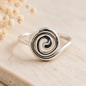 May include: A polished silver ring featuring a prominent spiral design on the face. The metal band is smooth and continuous, leading into a circular, coiled pattern that sits centered on the top of the ring, showcasing a reflective metallic finish.