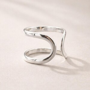 May include: A silver-colored ring with a unique, open design. The ring features a curved, double-band structure, creating an open space in the center. The polished metal reflects light, highlighting the smooth, flowing lines of the jewelry.