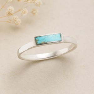May include: A silver ring featuring a rectangular, horizontal inlay of light blue turquoise stone. The band is smooth and polished, showcasing a clean, minimalist design with the vibrant blue stone set securely in the center of the metal frame.