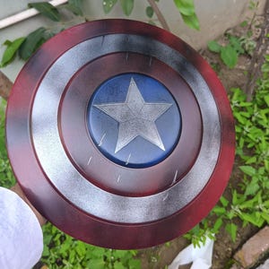 May include: A Captain America shield replica. The shield is circular, with a red outer ring, a silver inner ring, and a blue center with a silver star. The shield is held against a background of green foliage.