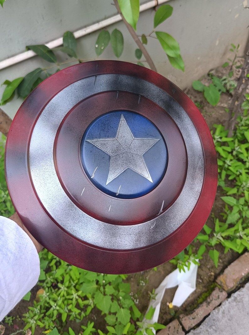 May include: A Captain America shield replica. The shield is circular, with a red outer ring, a silver inner ring, and a blue center with a silver star. The shield is held against a background of green foliage.