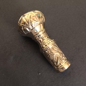 May include: A gold-colored, ornate walking cane handle. The handle features detailed floral and geometric patterns. The top is rounded with a textured design. The handle is made of metal and has a polished finish.