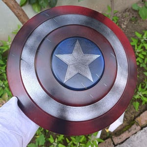 May include: A round shield with a red, white, and blue color scheme. The shield features a central blue circle with a white star, surrounded by concentric rings of silver, and red. The shield is a prop or collectible.