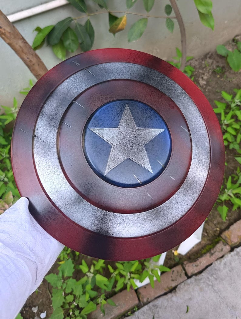 May include: A round shield with a red, white, and blue color scheme. The shield features a central blue circle with a white star, surrounded by concentric rings of silver, and red. The shield is a prop or collectible.