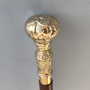 May include: A gold-colored walking cane with a decorative floral design on the handle and a dark brown wooden shaft. The handle has a rounded top with a detailed pattern. The cane is designed for support and style.