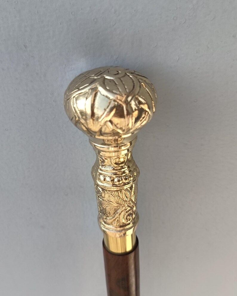 May include: A gold-colored walking cane with a decorative floral design on the handle and a dark brown wooden shaft. The handle has a rounded top with a detailed pattern. The cane is designed for support and style.