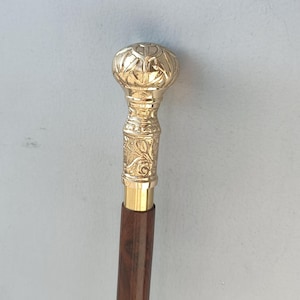 May include: A decorative walking cane with a gold-colored, ornate handle and a dark brown wooden shaft. The handle features detailed floral and geometric patterns. The cane is designed for support and style.