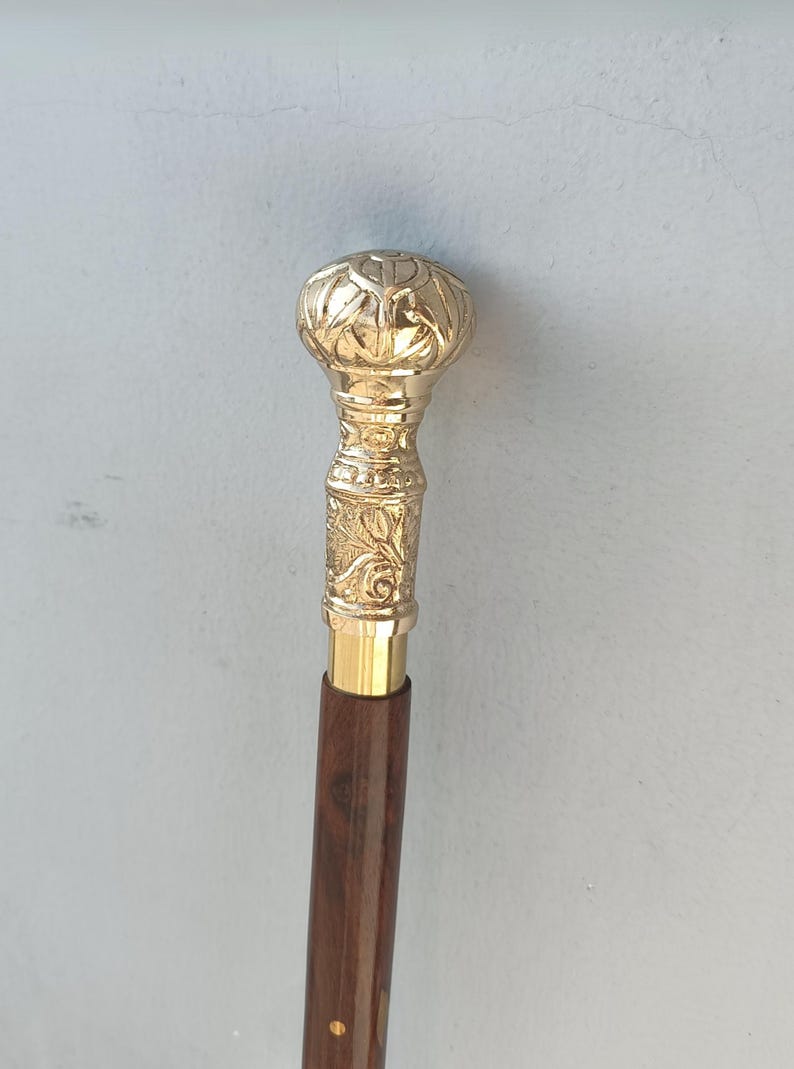 May include: A decorative walking cane with a gold-colored, ornate handle and a dark brown wooden shaft. The handle features detailed floral and geometric patterns. The cane is designed for support and style.