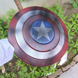 May include: A Captain America shield replica. The shield is round, with a red outer ring, a silver middle ring, and a blue center with a white star. The shield has a distressed, battle-worn appearance.