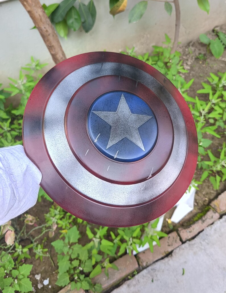 May include: A Captain America shield replica. The shield is round, with a red outer ring, a silver middle ring, and a blue center with a white star. The shield has a distressed, battle-worn appearance.
