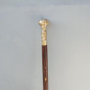 May include: A decorative walking cane with a gold-colored, ornate handle and a dark brown wooden shaft. The handle features detailed carvings and a rounded top. The shaft has gold-colored accents. The cane is approximately 36 inches long.