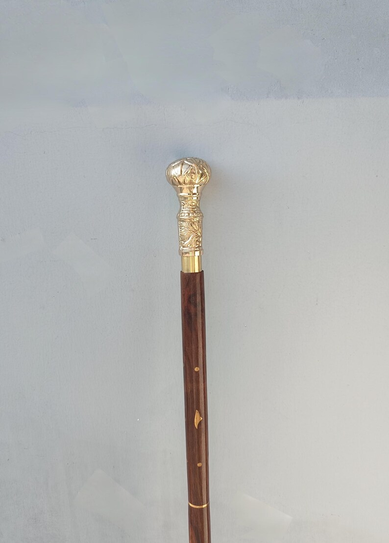 May include: A decorative walking cane with a gold-colored, ornate handle and a dark brown wooden shaft. The handle features detailed carvings and a rounded top. The shaft has gold-colored accents. The cane is approximately 36 inches long.
