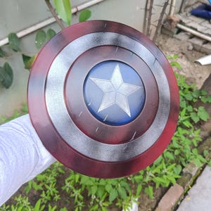 May include: A round shield with a red outer ring, a silver middle ring, and a blue center featuring a white star. The shield has a distressed, weathered appearance, suggesting it is a prop or replica.
