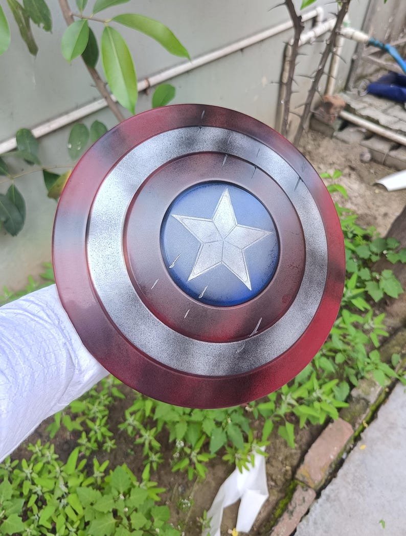 May include: A round shield with a red outer ring, a silver middle ring, and a blue center featuring a white star. The shield has a distressed, weathered appearance, suggesting it is a prop or replica.