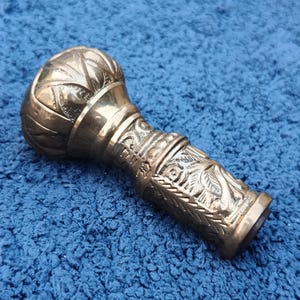 Könnte beinhalten: Ein detaillierter Messing-Stockgriff mit einem verzierten Design. Der Griff hat eine abgerundete Oberseite mit blattartigen Mustern, ein Mittelband mit eingravierten Details und einen unteren Abschnitt mit aufwendigen Schnitzereien. Das Messing hat eine polierte Oberfläche.