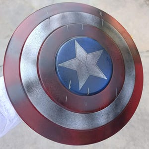 May include: A round shield with a red, silver, and blue color scheme. The shield features a central blue circle with a white star, surrounded by silver and red concentric rings. The shield has a distressed, battle-worn appearance.