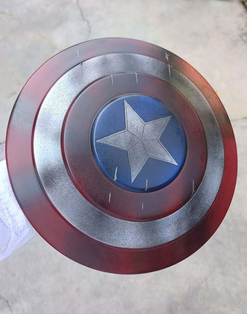 May include: A round shield with a red, silver, and blue color scheme. The shield features a central blue circle with a white star, surrounded by silver and red concentric rings. The shield has a distressed, battle-worn appearance.