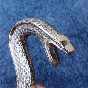 Puede incluir: Un mango de bastón plateado pulido con forma de cabeza de serpiente. La cabeza de la serpiente está detallada con escamas y un patrón texturizado. El mango del bastón es curvo y tiene la boca abierta.