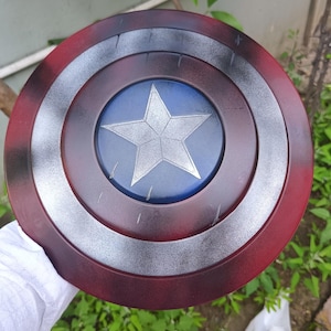 May include: A round shield with a red, silver, and blue color scheme. The shield features a central blue circle with a silver star, surrounded by concentric rings of silver, black, and red. The shield is held by a person.