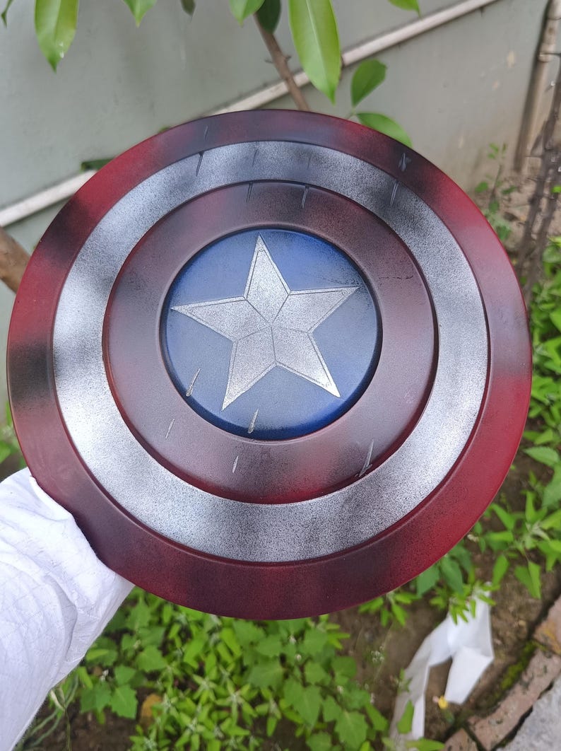 May include: A round shield with a red, silver, and blue color scheme. The shield features a central blue circle with a silver star, surrounded by concentric rings of silver, black, and red. The shield is held by a person.
