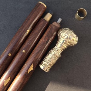 May include: A disassembled wooden walking cane with a brass handle and accents. The dark brown wooden shaft is in three sections, with gold-colored metal details. The handle is ornate, with a bulbous top and floral carvings.